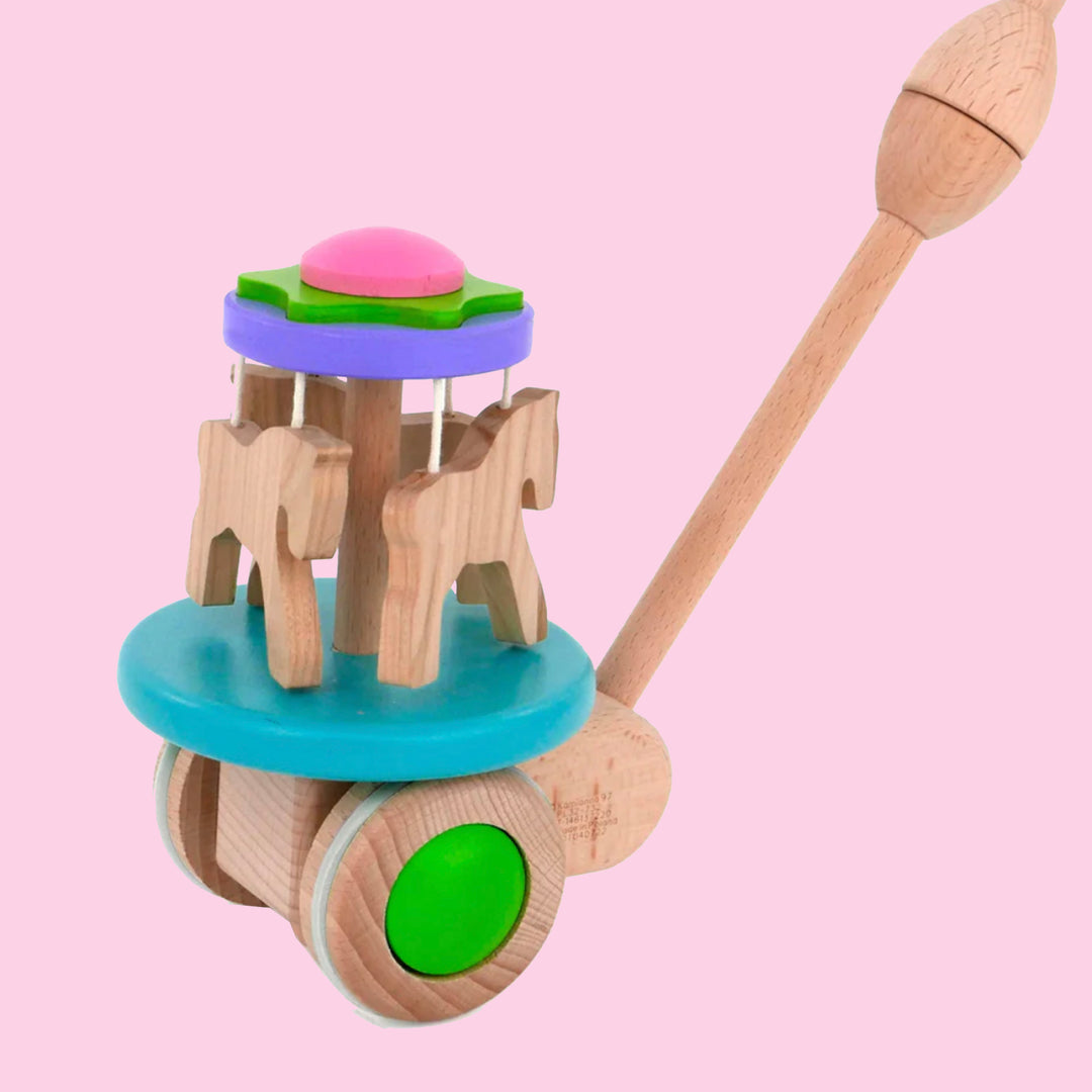 Wooden carousel push toy featuring wooden horses on blue platform and green and pale wood wheels. on light pink background.