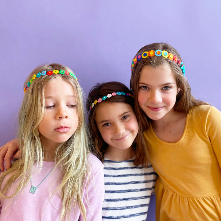 Three kids wearing plastic flower crowns. They are in front of a purple background.