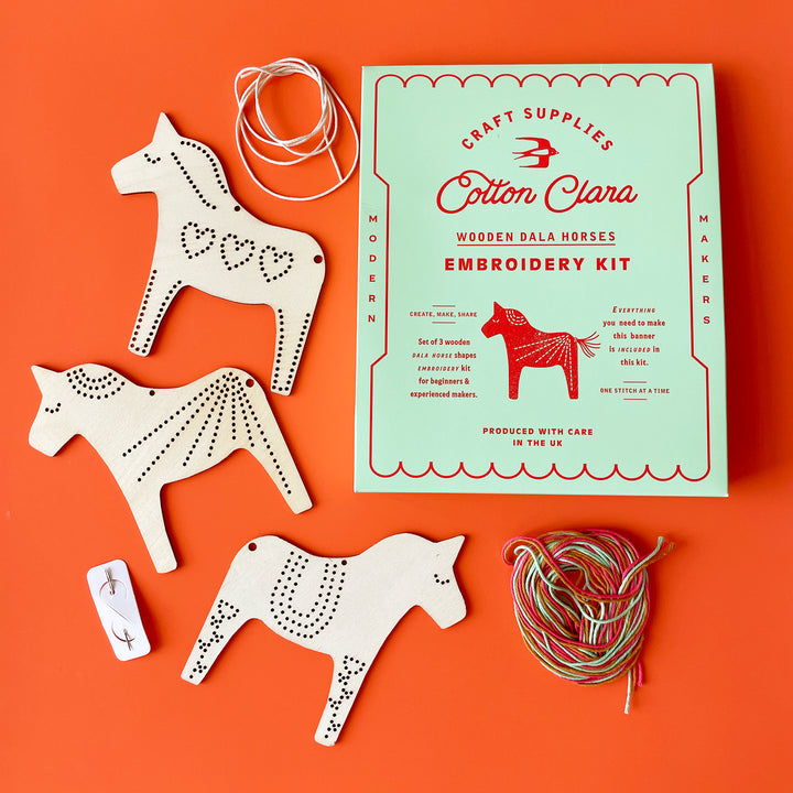 The contents of a Dala House embroidery kit laying flat: Three flat wooden horse shapes, a light green box, and two piles of string.