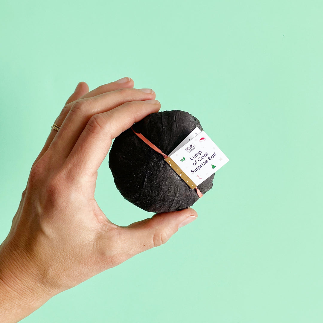 A hand holding a black lump of coal-inspired surprise bal with speckles of glitter and a small tag attached with an orange band.