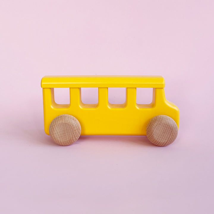 Simple wooden school bus with yellow body and pale wood colored wheels. Light pink background.