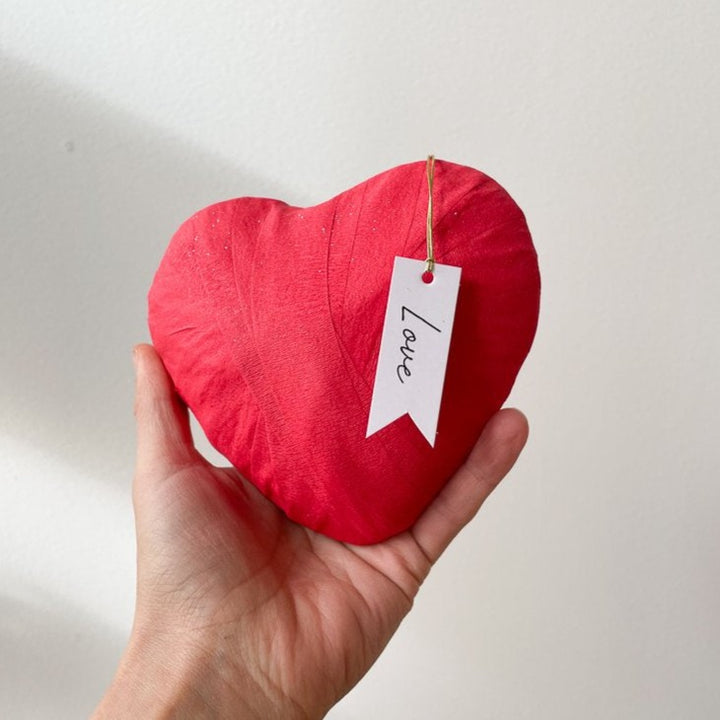 A hand holding a large, red, heart-shaped surprise ball with a tag reading "Love" in a script handwriting.