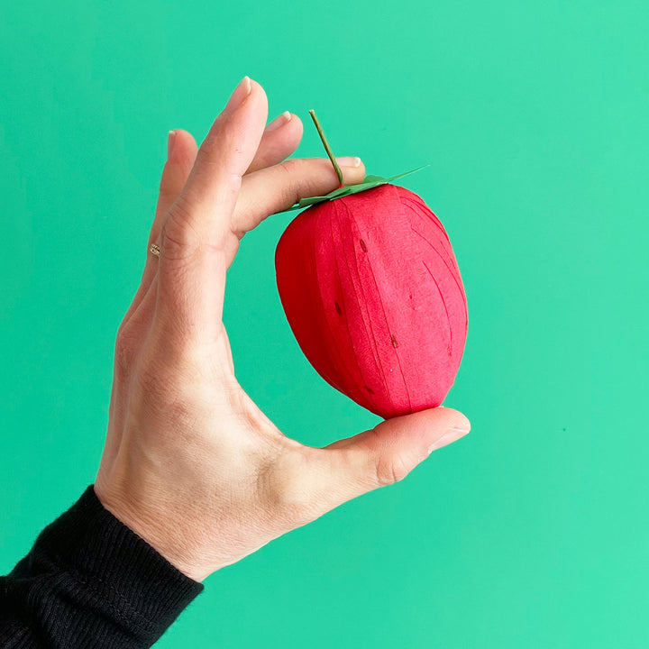 A hand holding a strawberry-shaped srprise ball against a bright green background.