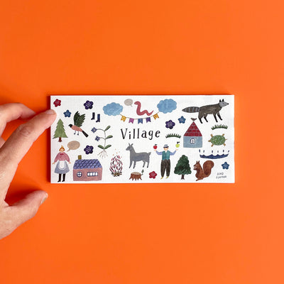 The Village memo pad by Aiko Fukawa on an orange background, with a hand for scale.
