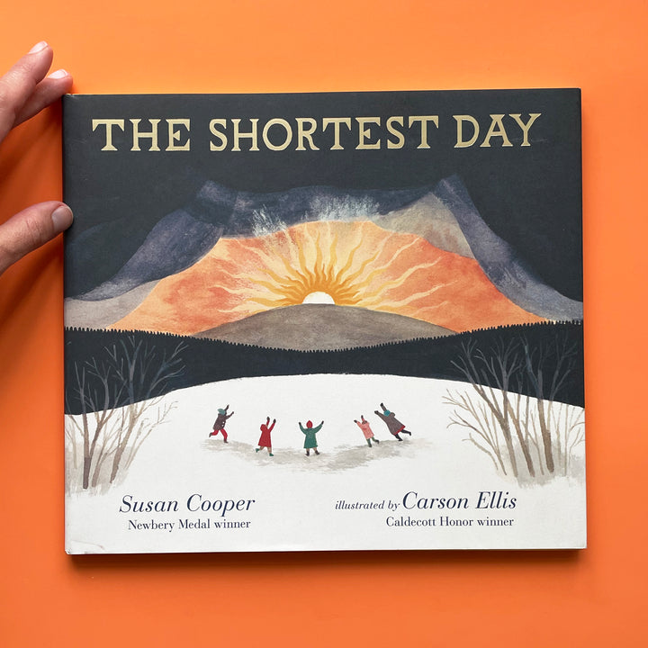 The Shortest Day bok against an orange background. The book cover features a watercolor illustration of a bright orange sunset beyond a dark hillside. Five small figure rejoice in a the faoreground. 