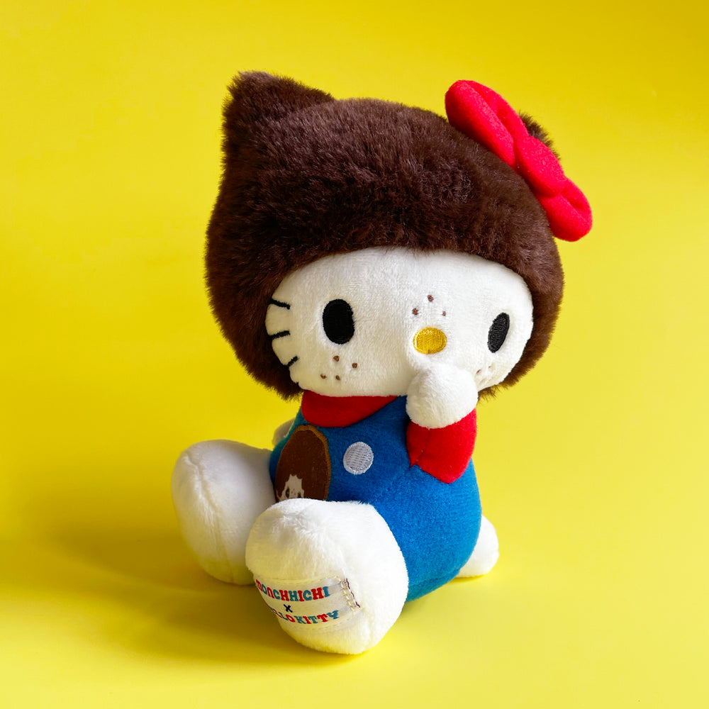 Plush toy resembling Hello Kitty wearing a  brown fur hood with red bow, and a blue outfit. Sitting on a yellow background. 