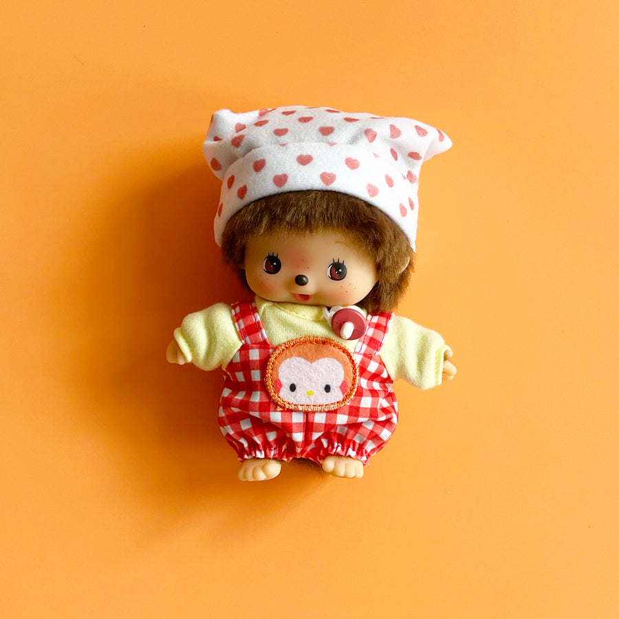 Monchhichi doll wearing a red checkered outfit with a white heart pattern on an orange background