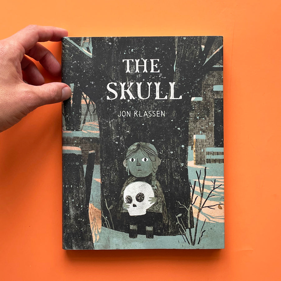 The Skull book displayed on an orange background. The cover features an illustration of a young girl with wide eyes standing against a tree in the snow. In her hands, she holds a bright white skull. 