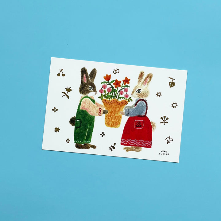 A postcard with an illustration of two anthropomorphic rabbits in outfits holding a bouquet of flowers on a white background.