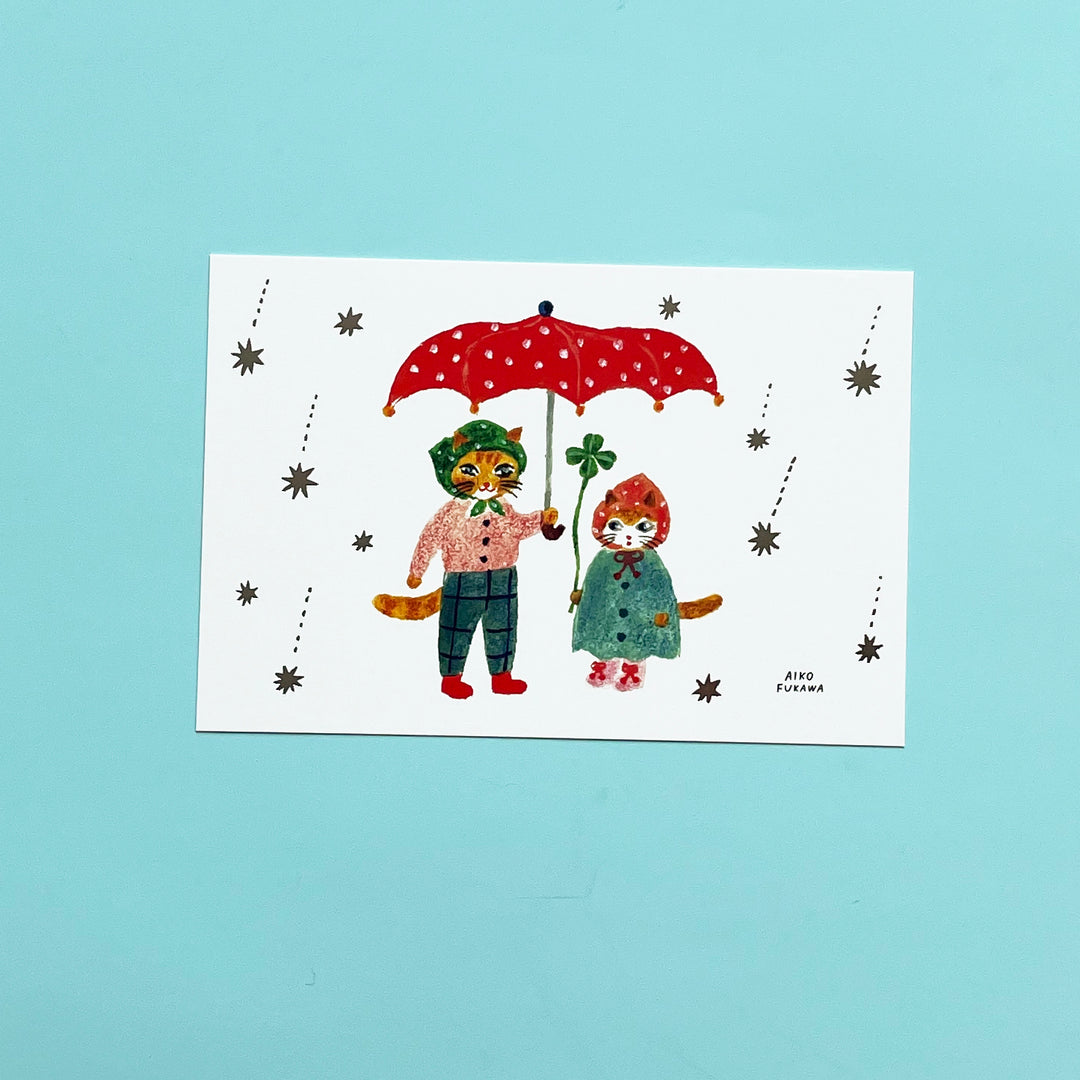 A post card with two anthropomorphic cats in outfits sharing a red umbrella as starts fall around them.