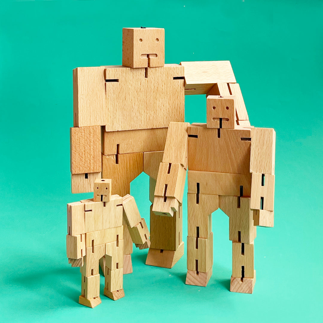 Three robot shaped wooden toys in small medium and large sizes standing up against a bright green background. The toys are a light natural wood color. 