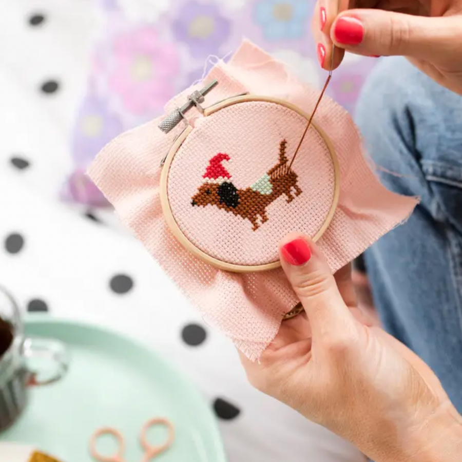 Hands working on a cross stitch project featuring a dachshund wearing a Santa hat and small green blanket. 