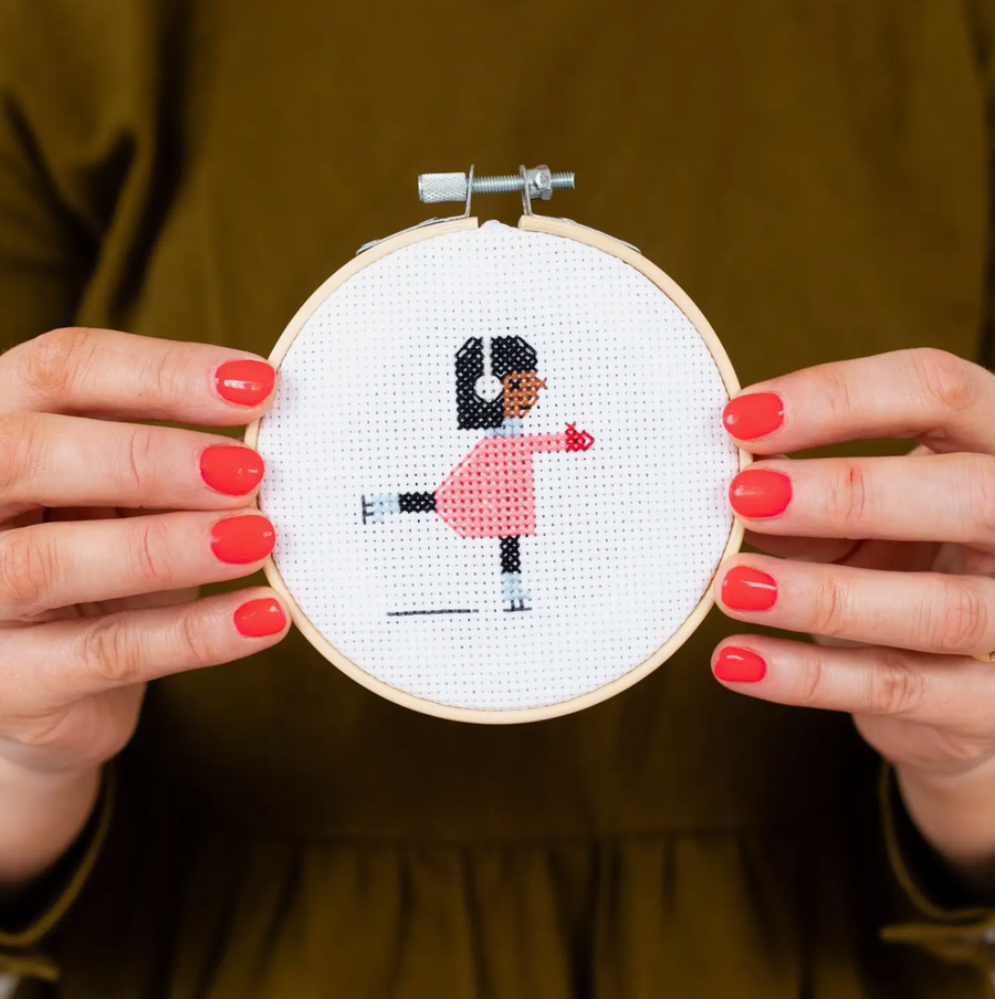 Hands holding a small hoop with a finished cross stitch project. Embroidered image is of a young girl ice skating in a pink coat, red mittens and white ear muffs. 