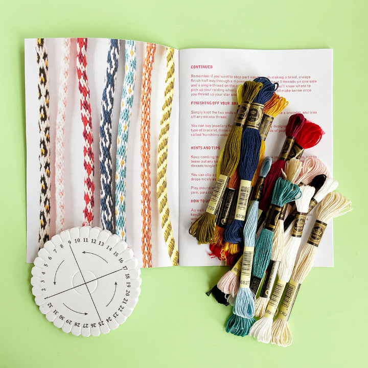Pattern book for Kumihimo cords open to a photograph of finihed decorative cords. A colorful pile of embroidery floww is scattered across the book and a Kumihimo tool is set to the left. 