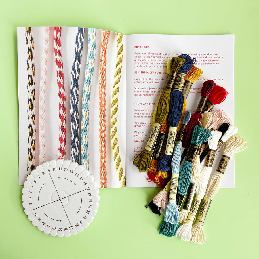 Pattern book for Kumihimo cords open to a photograph of finihed decorative cords. A colorful pile of embroidery floww is scattered across the book and a Kumihimo tool is set to the left. 
