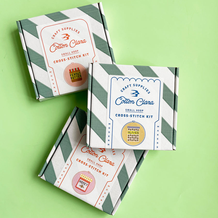 A scattered stack of three cross stitch kits in bold, striped boxes. The kits are for a cross stitch jar of pickles, jar of olives and jar of honey. 