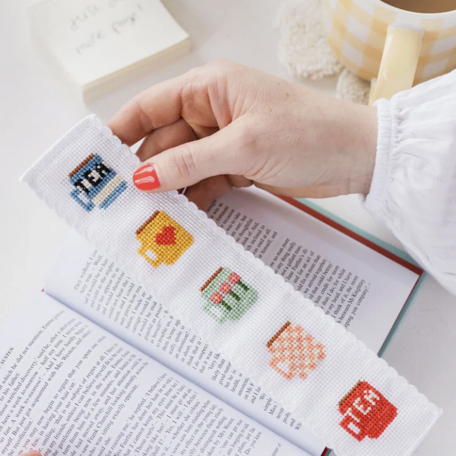 A hand holding a cross stitched book mark featuring five mugs with different, colorful patterns. 