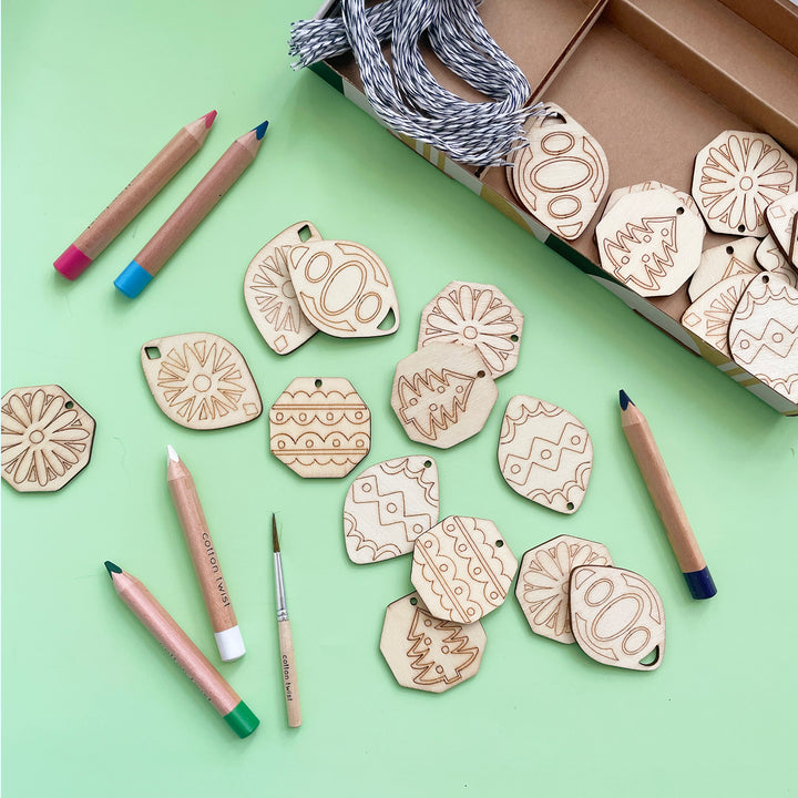 A Paint Your Own Wooden Ornament kit open and displaying what's inside including several unpainted wooden ornaments, colored pencils, string and a paint brush. 