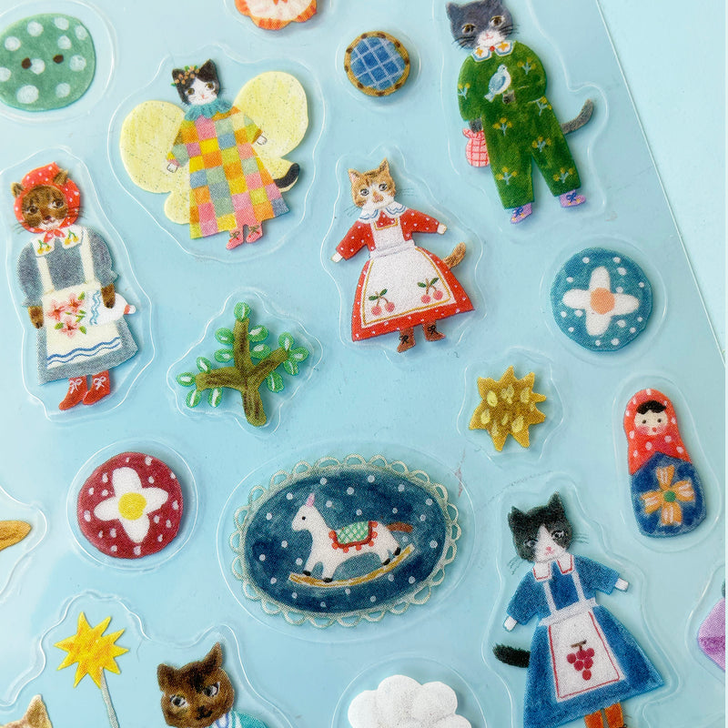 A closeup of the Cats sticker sheet featuring a grey cat in a green jumpsuit holding a bird, a tabby cat in a red dress and cherry apron, and a black and white cat in a large patterned dress with butterfly wings.