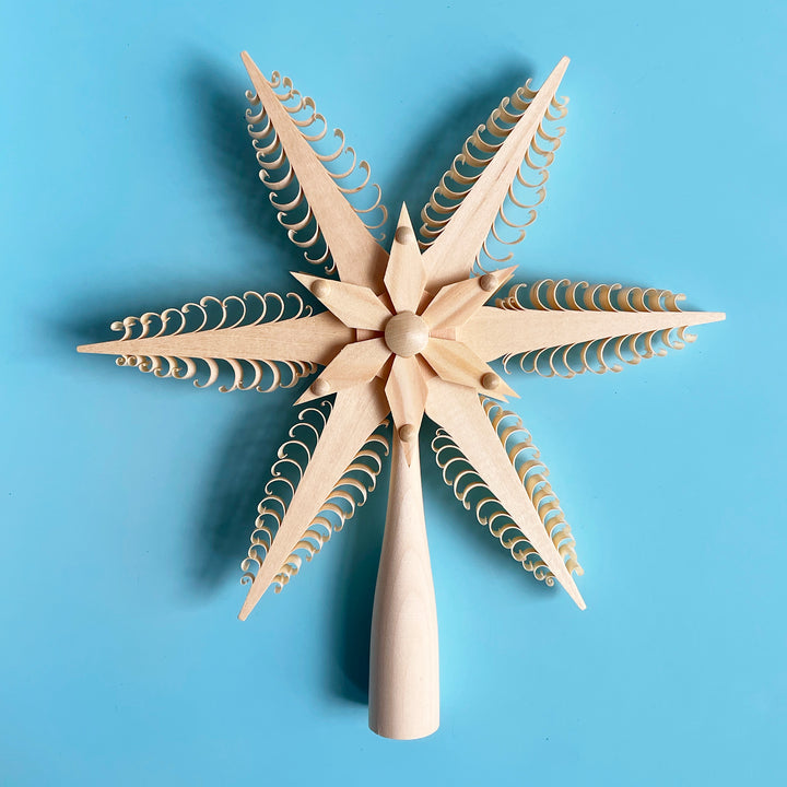 An intricate, natural carved wood star tree topper. The star features 6 points with a shaved, scallop design. 