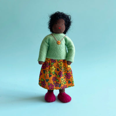 A 5 inch cotton doll stands wearing a mint felt top and an orange skirt with pink and yellow flowers. The doll has dark skin and black curly hair.