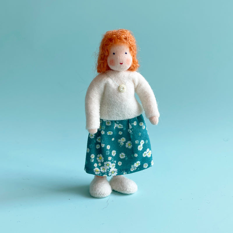 A 5 inch cotton doll stands wearing a white felt top and a blue skirt with white flowers. The doll has light skin and orange hair.