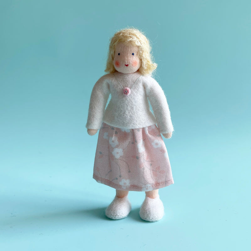 A 5 inch cotton doll stands wearing a white felt top and a pastel pink skirt with white flowers. The doll has light skin and blonde hair.