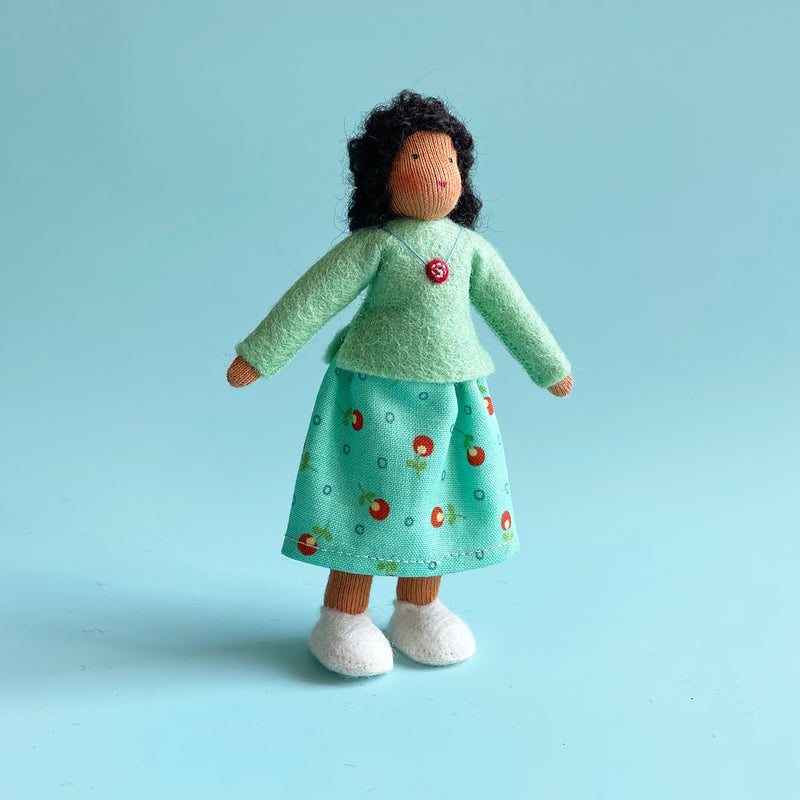 A 5 inch cotton doll stands wearing a mint felt top and a blue skirt with red flowers. The doll has medium skin and dark curly hair.