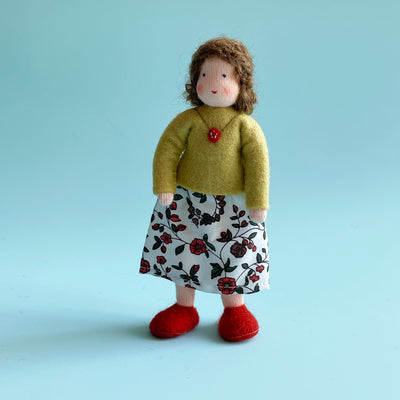 A 5 inch cotton doll stands wearing a green felt top and a white skirt with red flowers and green leaves. The doll has light skin and brown hair.
