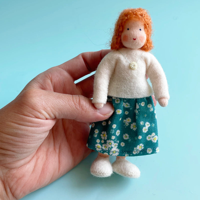 A hand holds a 5 inch cotton doll wearing a white felt top and a blue skirt with white flowers. The doll has light skin and orange hair.