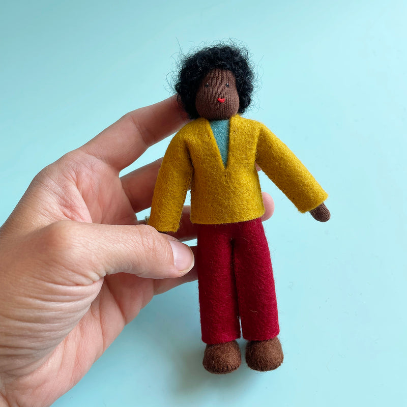A hand holds a 5.25-inch cotton doll with dark skin and black curly hair wearing a yellow felt top and yellow felt pants.