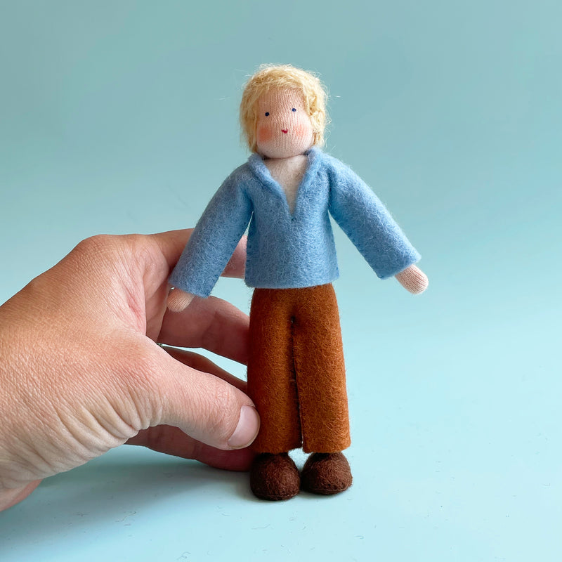 A hand holds a 5.25-inch cotton doll with light skin and blonde hair wearing a light blue felt top and brown felt pants.