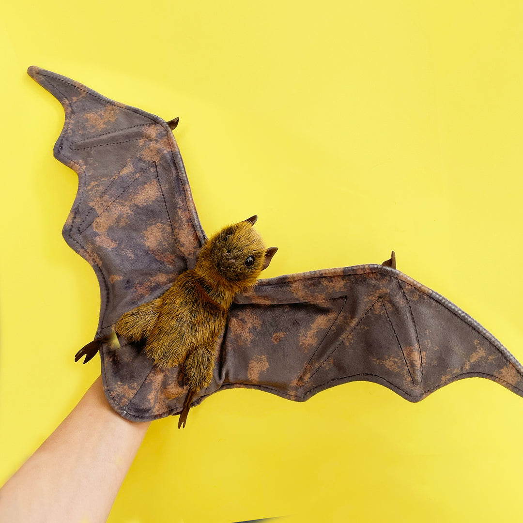 A fruit bat hand puppet with outstretched, mottled brown wings held against a bright yellow backdrop. 