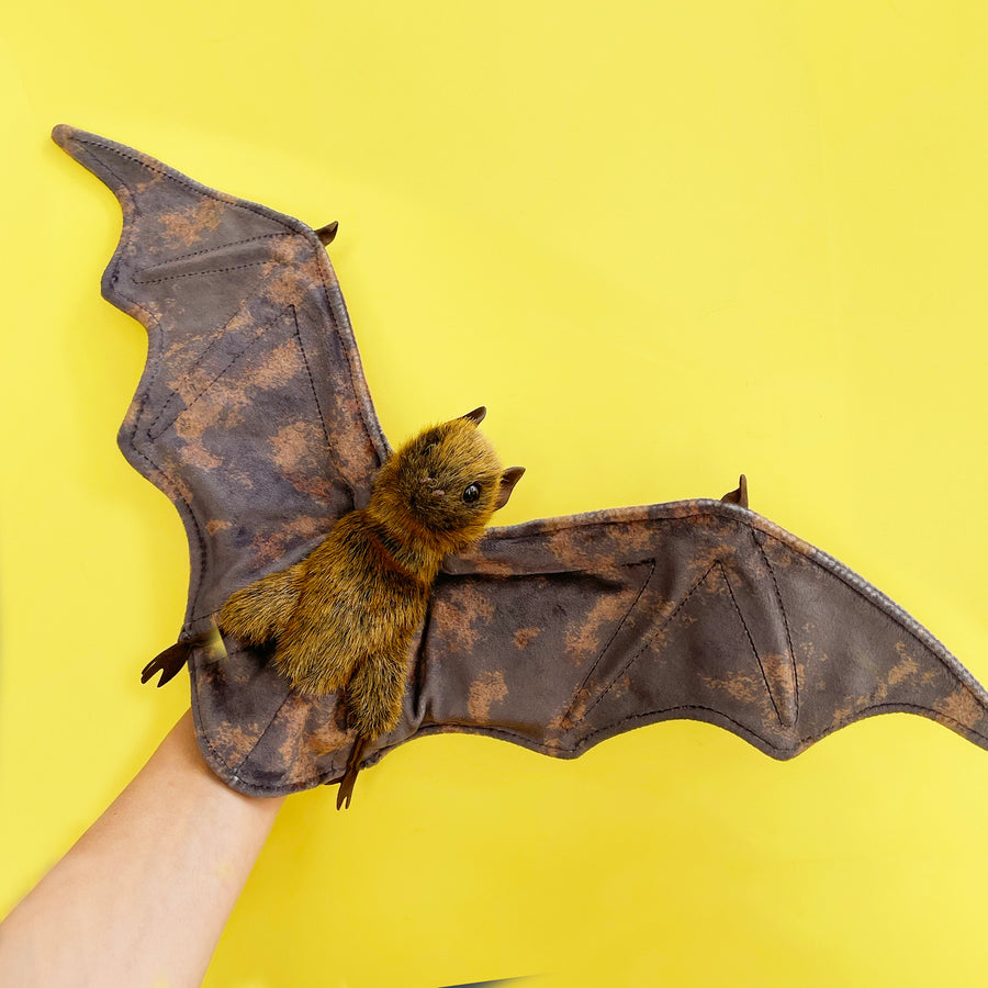 A fruit bat hand puppet with outstretched, mottled brown wings held against a bright yellow backdrop. 