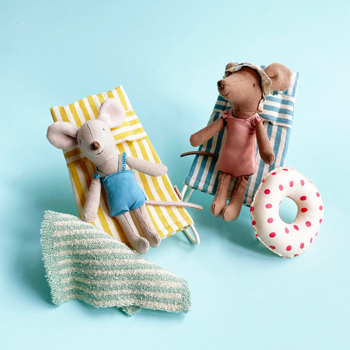 Two miniature fabric mice relax on striped beach chairs surrounded by beach accessories, including a tiny towel and polka-dot toy floatie. 