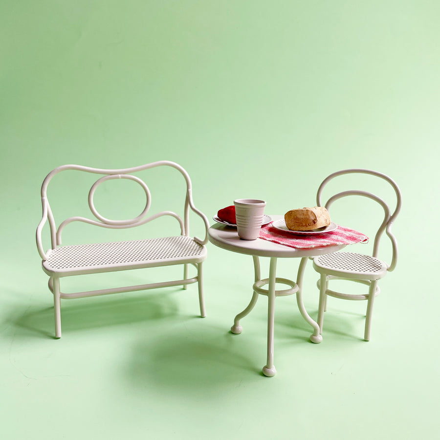 A miniature, white metal cafe table and chairs st up with a red gingham cloth, white table ware and a slice of cheese displayed on a light green background. 