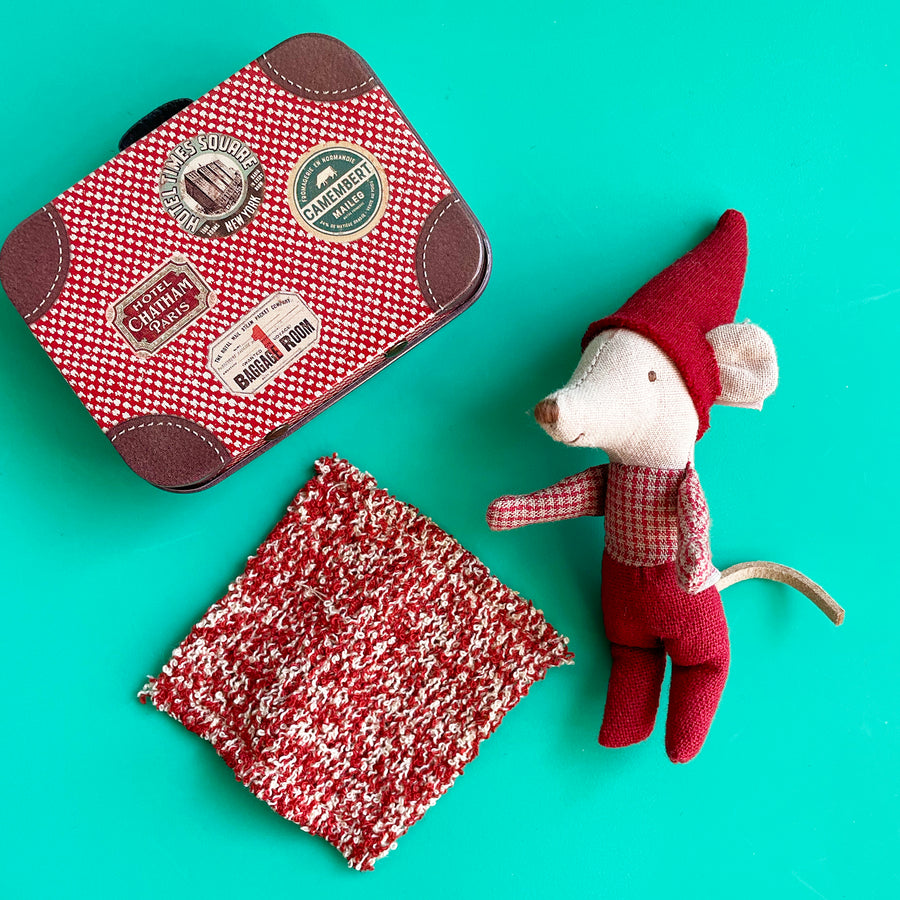 A miniature fabric mouse dressed up to look like a Christmas elf, displayed with a red and wite knit blanket and a red and white metal suitcase against a bright green background. 