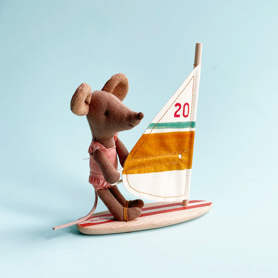 A miniature fabric toy mouse wearing a pink swimsuit rides a tiny wind sail against a light blue background. 