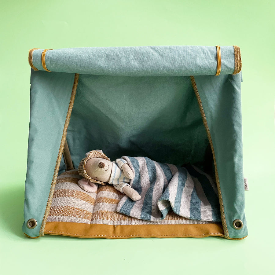 A miniature fabric toy mouse sleeps under a striped blanket inside a small green fabric tent against a light green background. 