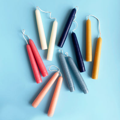 6 different colored pairs of 6 inch taper candles. Each pair is connected by the wicks. The candles are bright pink, light peach, white, light blue, dark blue, and natural beeswax.