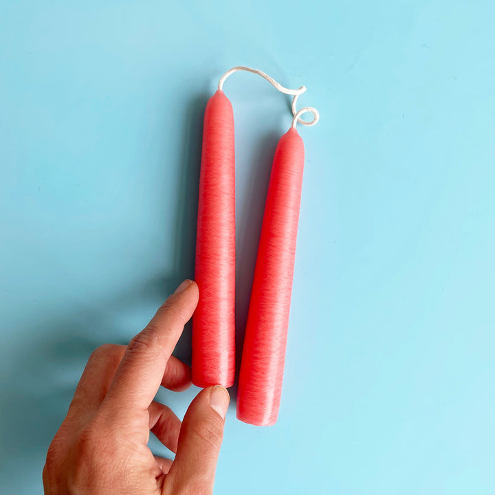 A pair of bright pink 6 inch taper candles connected by their wicks. A hand is also shown as a size reference.