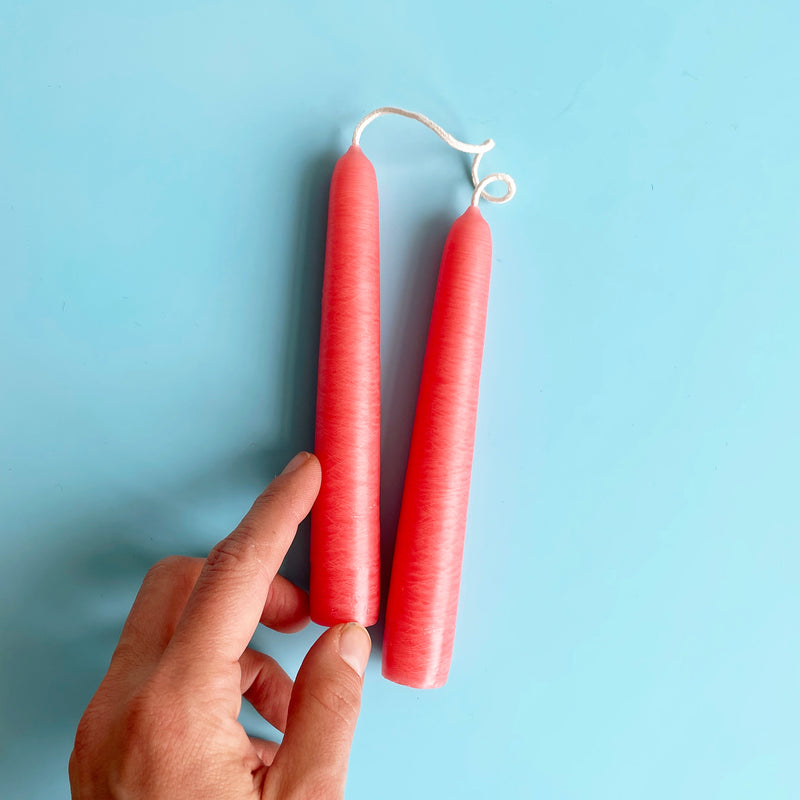 A pair of bright pink 6 inch taper candles connected by their wicks. A hand is also shown as a size reference.