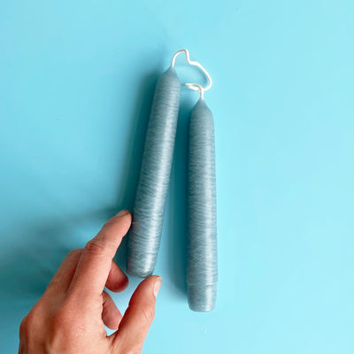 A pair of light blue 6 inch taper candles connected by their wicks, with a hand in frame for size reference.