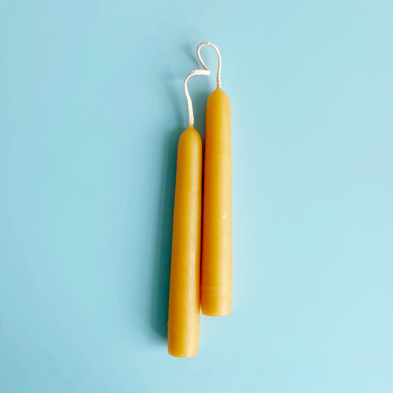 A pair of natural beeswax 6 inch taper candles connected by their wicks.