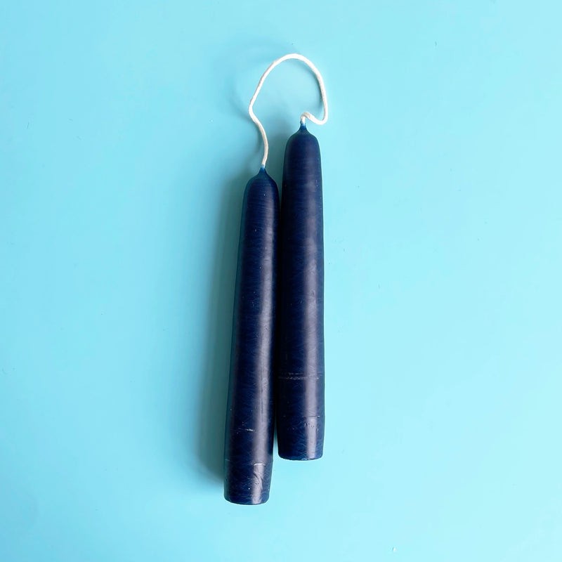 A pair of dark blue 6 inch taper candles connected by their wicks.