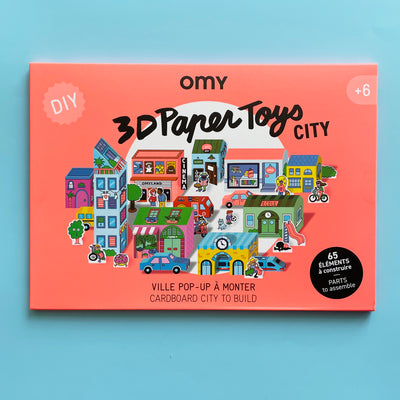 The packaging of the 3d paper city kit showing 3d paper buildings, characters, and vehicles in a cartoon art style. The package says "OMY DIY 3D Paper Toys city; 65 parts to assemble; cardboard city to build."