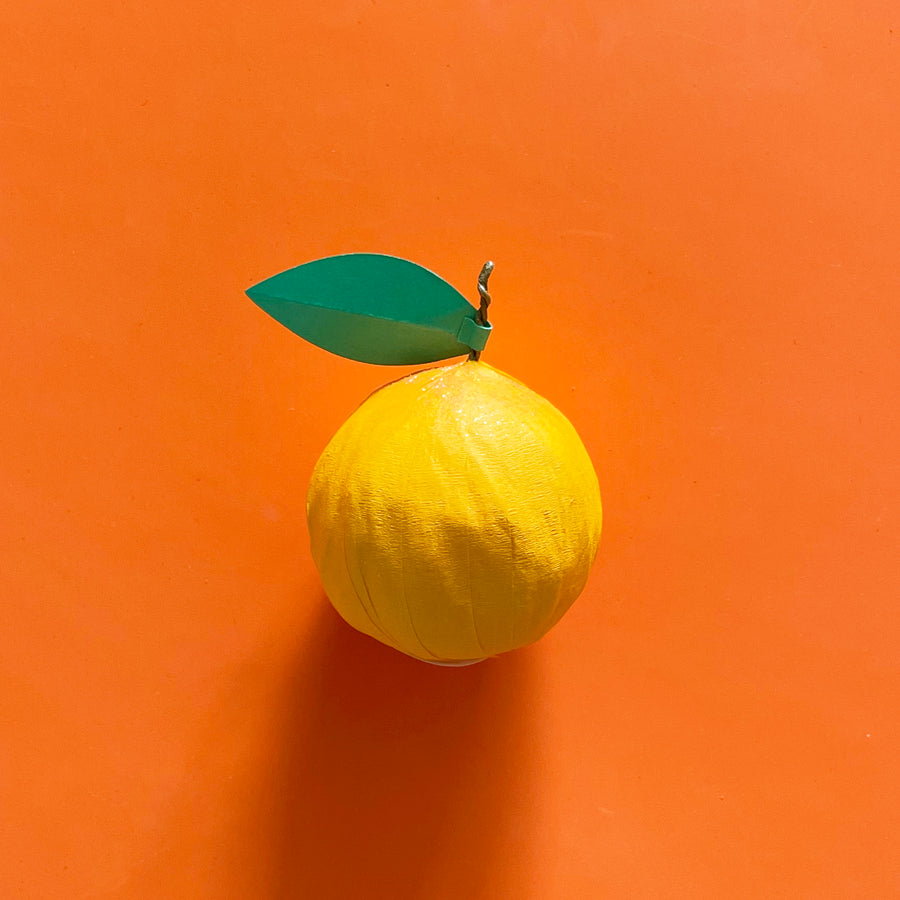 A golden yellow clenmentine-shaped surprise ball with a wire stem and green paper leaf against a bright orange background. 
