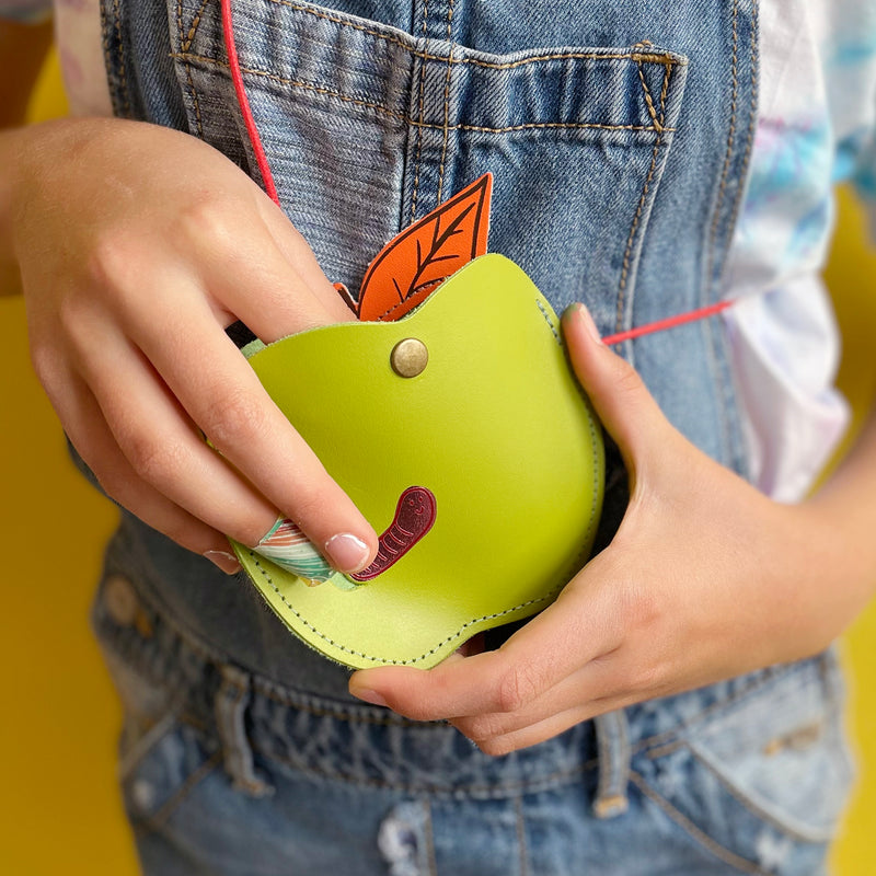 A green apple shaped purse being worn and held by a child wearing overalls