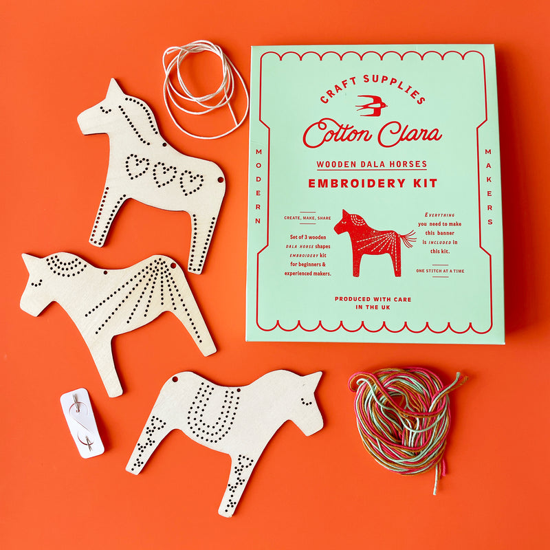 The contents of a Dala House embroidery kit laying flat: Three flat wooden horse shapes, a light green box, and two piles of string. 
