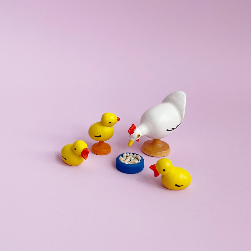 Wooden chicken figurine with three yellow checks around a blue bowl of food on a pink background. 
